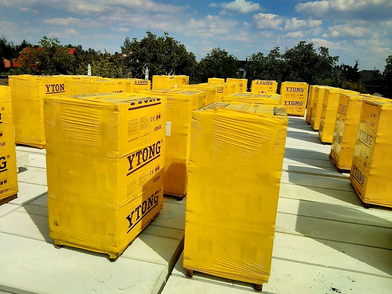 Palety z żółtą folią YTONG, ułożone na szarych płytach stropowych, widok z góry na tle zieleni i błękitnego nieba z chmurami.