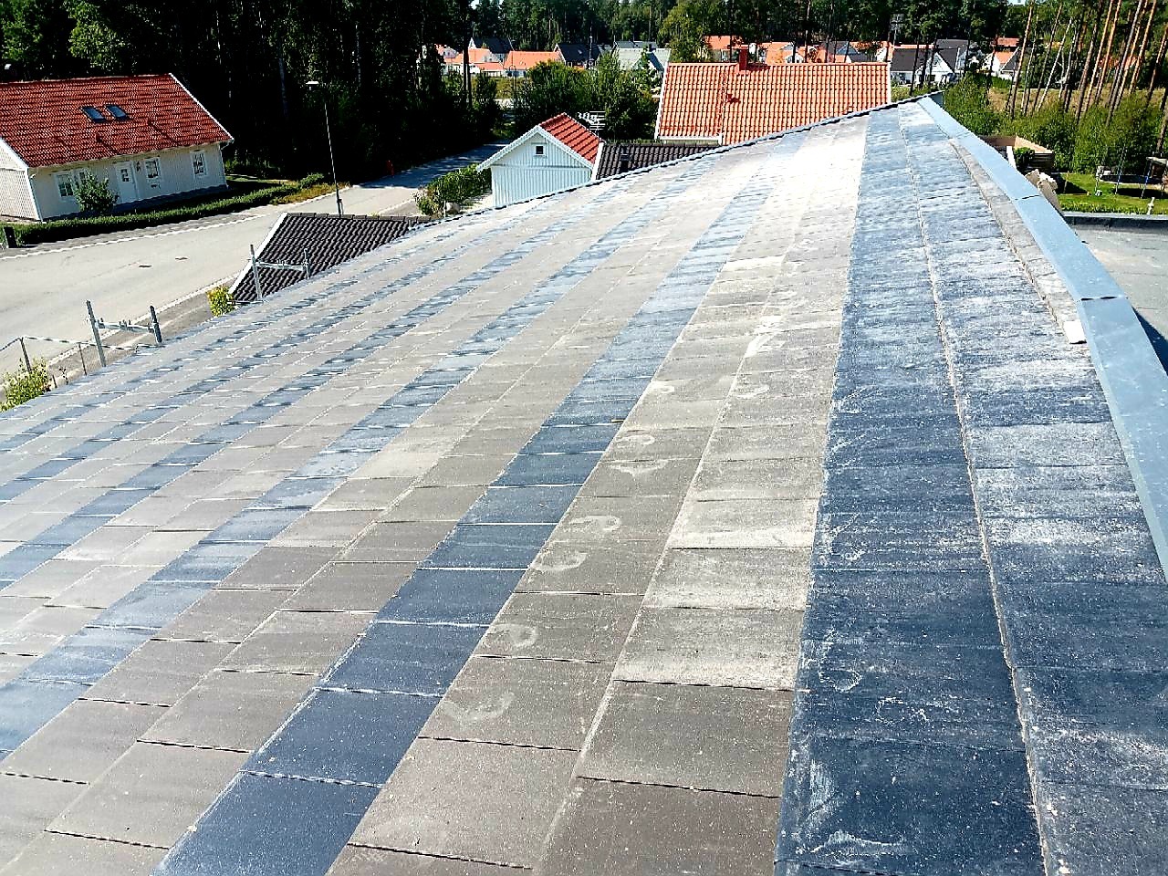 Płaski dach pokryty szarą kostką brukową, widok z góry na krawędź dachu z metalowym obrzeżem, w tle zabudowa jednorodzinna.