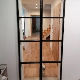 Przeszklone, dwuskrzydłowe drzwi przesuwne z czarnymi szprosami, widok na wnętrze domu z drewnianą podłogą i schodami, mały piesek w tle.