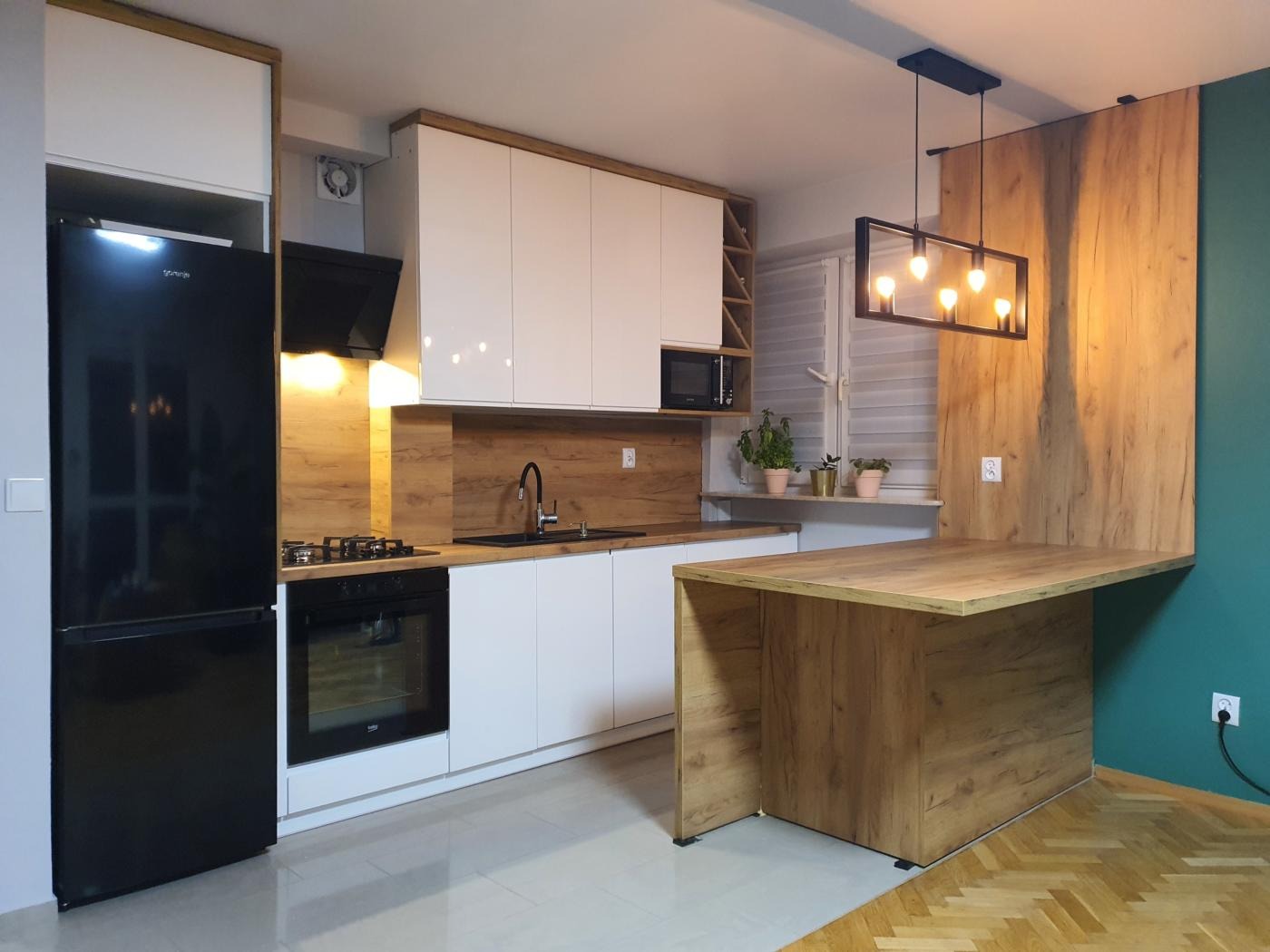 Nowoczesna kuchnia z białymi szafkami, drewnianym blatem i wyspą, czarnym sprzętem AGD i oświetleniem w stylu industrialnym.