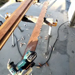 Deska z wyciętymi ozdobnymi krawędziami, leżąca na kozłach, obok akumulatorowa wyrzynarka Makita, w tle deski i betonowa posadzka z resztkami drewna.