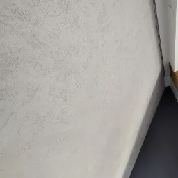 Jasnoszara ściana z dekoracyjną strukturą przypominającą przecierkę, oświetlona z boku, z widocznym fragmentem czarnej podłogi i białego narożnika sufitu z listwą.