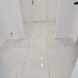 Białe płytki podłogowe imitujące marmur w korytarzu z białą listwą przypodłogową i drzwiami z złotą klamką.