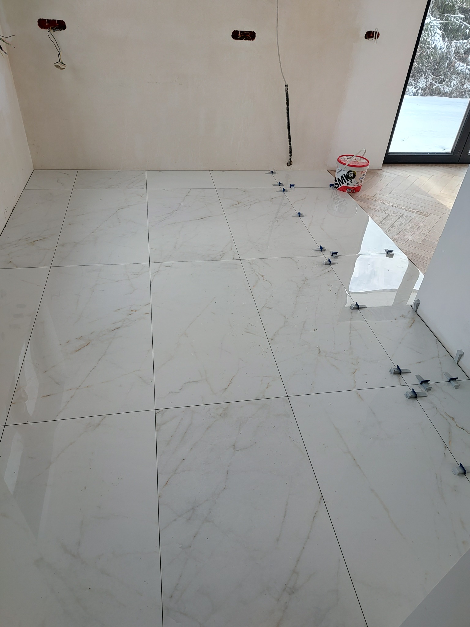 Układanie białych, marmurkowych płytek podłogowych z użyciem niebieskich krzyżyków dystansowych; wiaderko z klejem w tle, widoczne niedokończone instalacje elektryczne w ścianie.