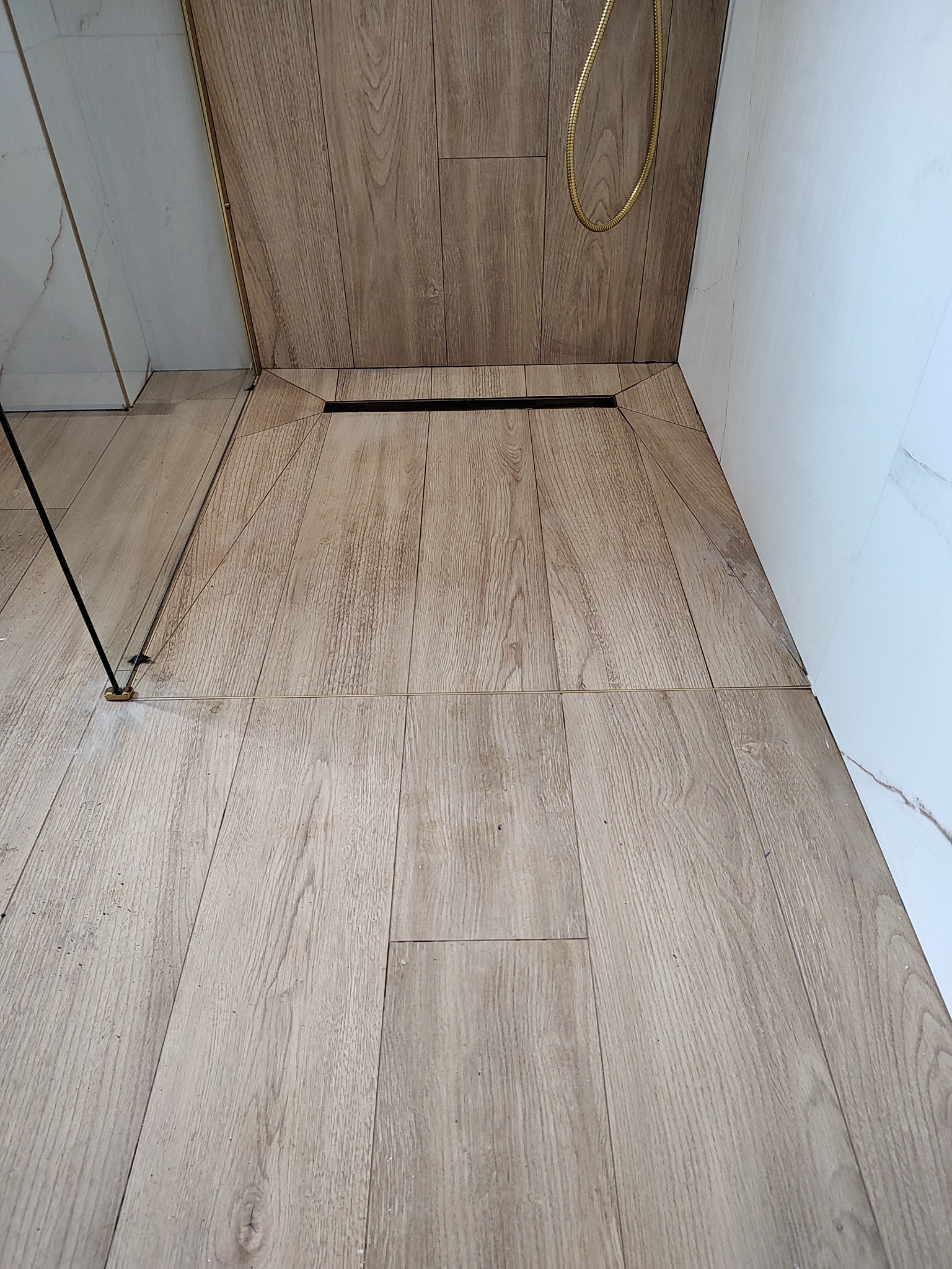 Ukończona łazienka z odpływem liniowym w podłodze wyłożonej płytkami imitującymi drewno, kabina prysznicowa ze złotymi elementami oraz białe płytki na ścianie.