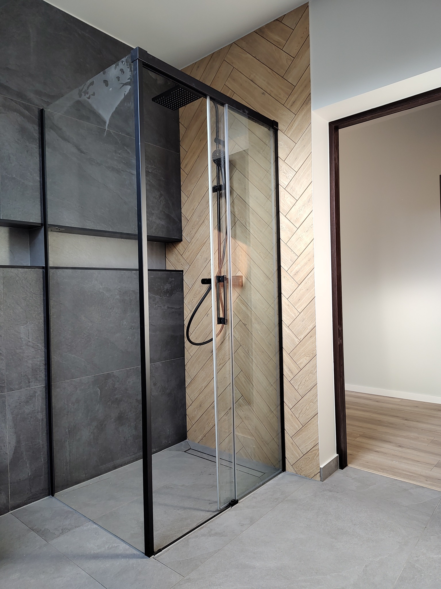 Nowoczesna kabina prysznicowa z czarną ramą, szklaną ścianą i deszczownicą, z widocznymi plamami wody na szkle, obok ściana z płytek ułożonych w jodełkę i otwarte drzwi do pokoju z podłogą...
