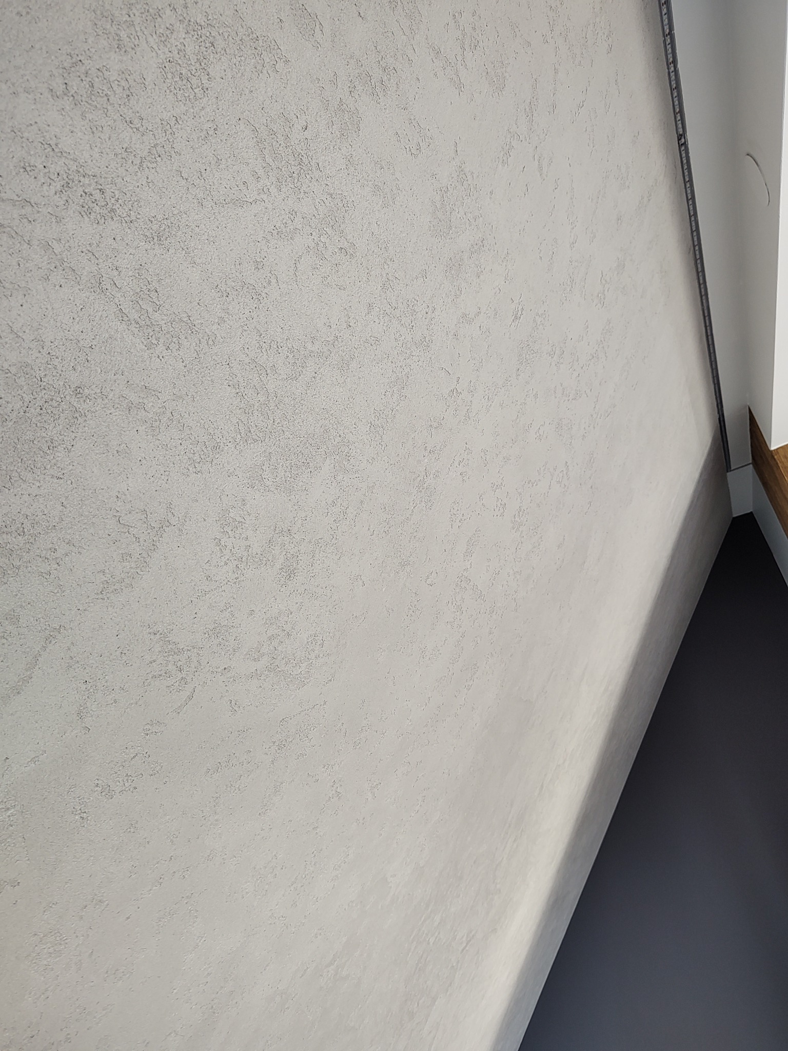 Jasnoszara ściana z dekoracyjną strukturą przypominającą przecierkę, oświetlona z boku, z widocznym fragmentem czarnej podłogi i białego narożnika sufitu z listwą.