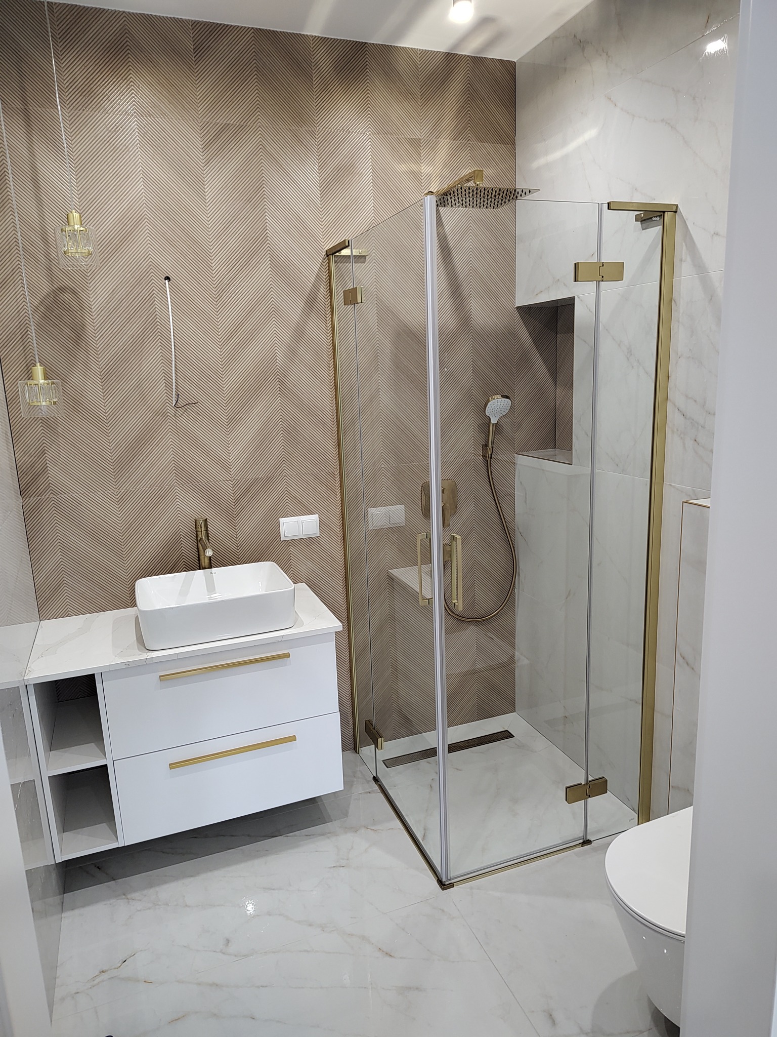 Nowoczesna łazienka z kabiną prysznicową ze złotymi detalami, białą umywalką nablatową na szafce z marmurowym blatem i chevronową ścianą akcentującą