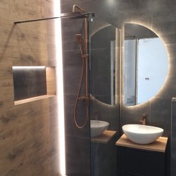 Nowoczesna łazienka z kabiną typu walk-in, drewnopodobnymi i szarymi płytkami, okrągłym lustrem z podświetleniem LED i złotą armaturą. Wnęka w ścianie z podświetleniem.