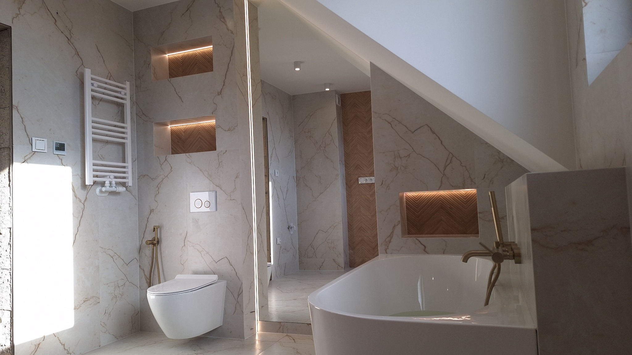 Elegancka łazienka na poddaszu z marmurowymi płytkami, podświetlanymi niszami i białą armaturą. Widoczny grzejnik drabinkowy, toaleta i wanna z złotą baterią.