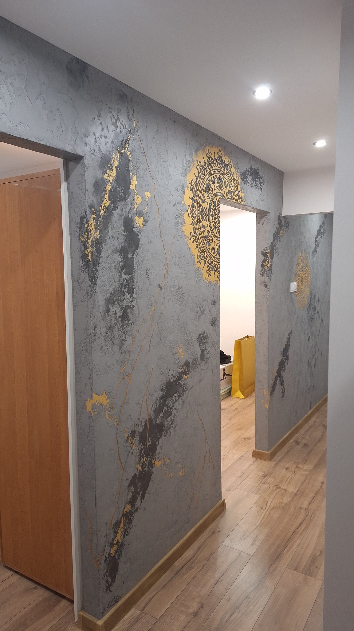 Korytarz z szarą ścianą zdobioną złotymi wzorami i mandalą, drewniana podłoga, widok na kolejne pomieszczenie. Dekoracyjne wykończenie wnętrza.
