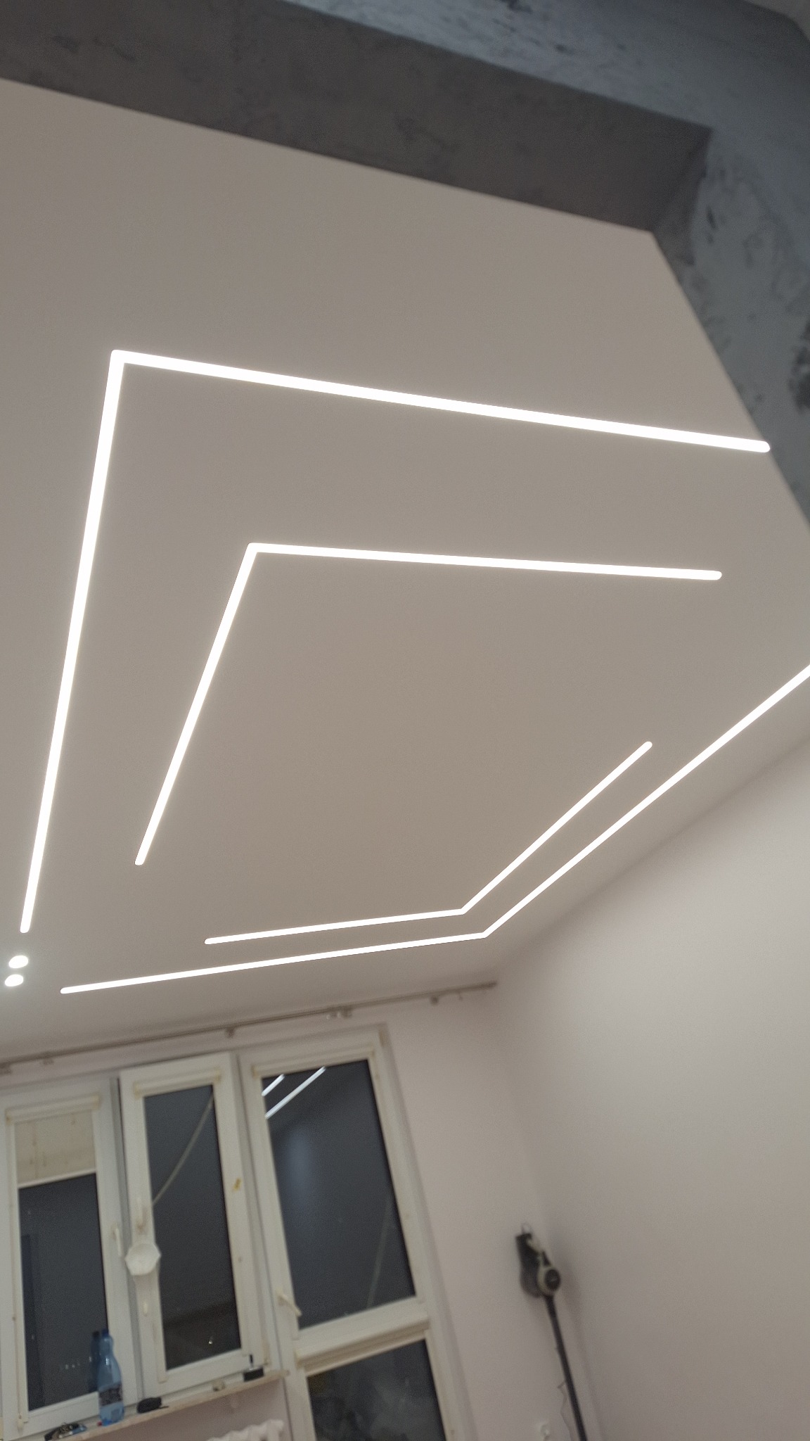 Nowoczesny sufit z liniowym oświetleniem LED w kształcie geometrycznym, widok z dołu, okno w tle, minimalistyczny design.