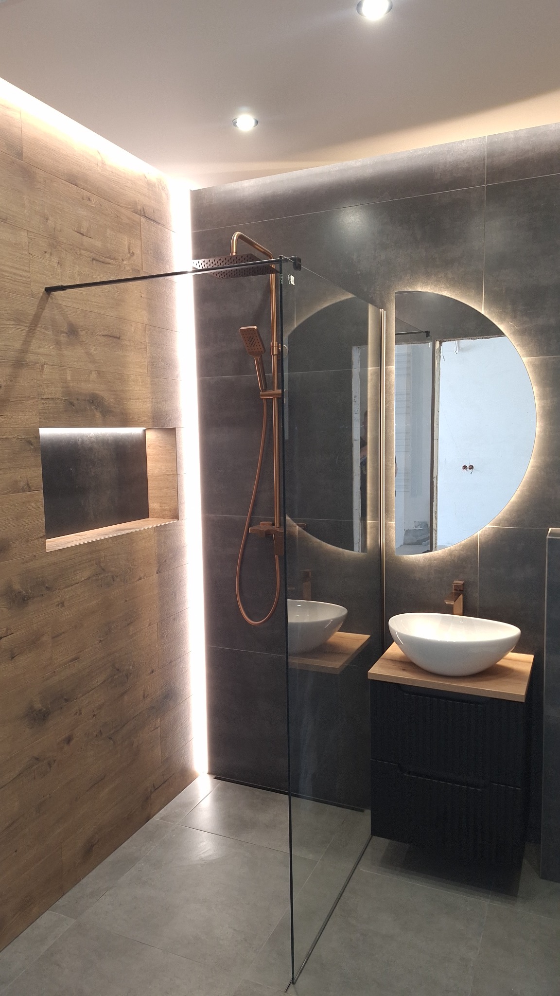 Nowoczesna łazienka z kabiną typu walk-in, drewnopodobnymi i szarymi płytkami, okrągłym lustrem z podświetleniem LED i złotą armaturą. Wnęka w ścianie z podświetleniem.