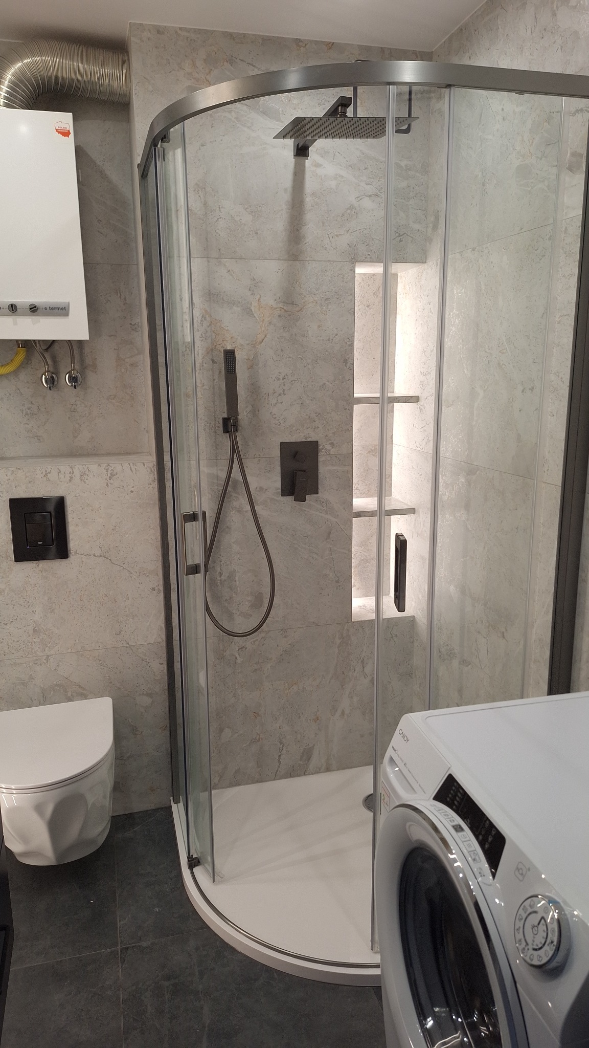 Nowoczesna kabina prysznicowa z deszczownicą i półkami wnękowymi, obok toaleta i pralka. Płytki imitujące marmur. Grzejnik nad toaletą.
