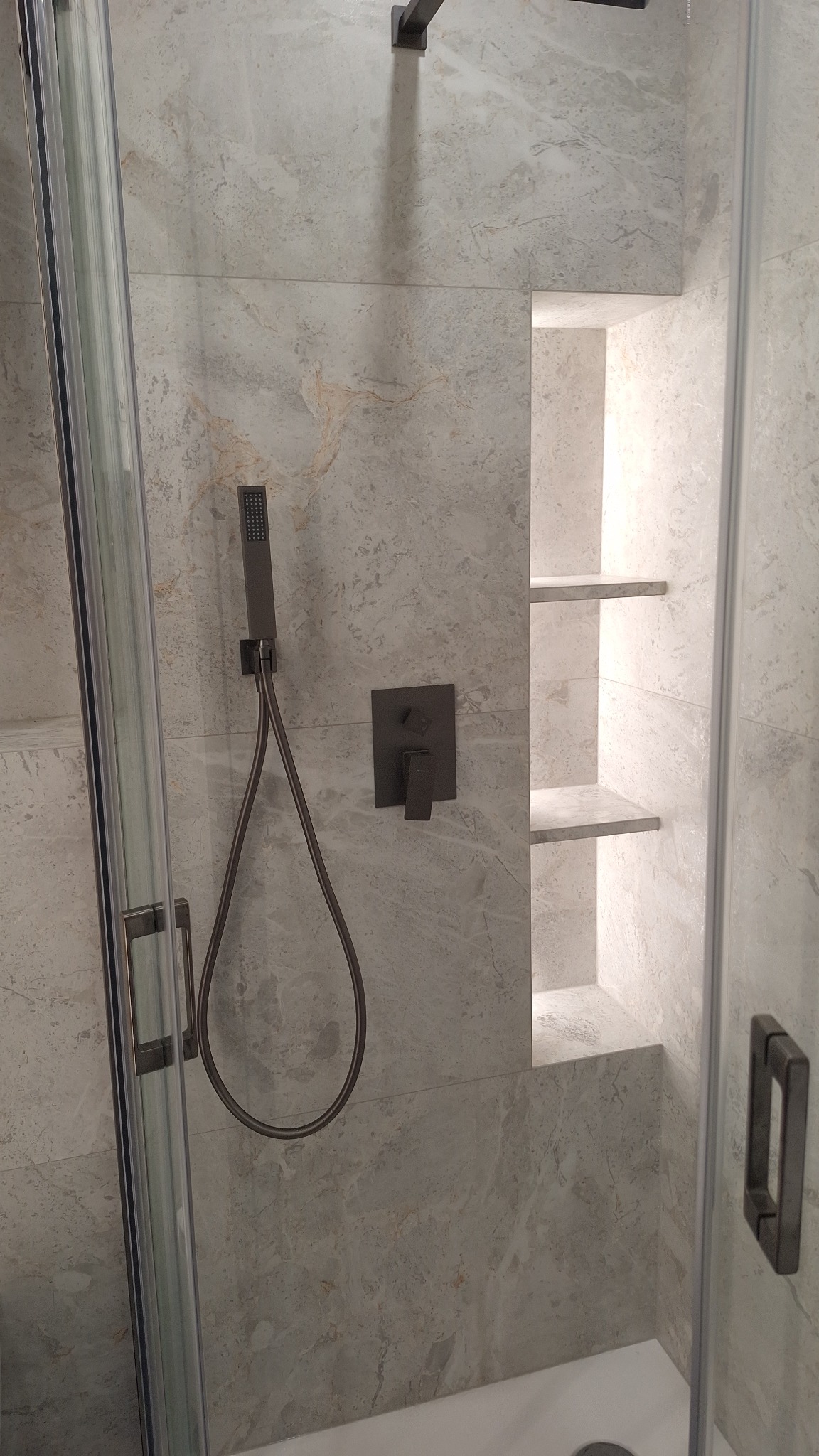 Nowoczesna kabina prysznicowa z szarymi płytkami imitującymi marmur, wnęką z półkami i czarną armaturą. Minimalistyczny design, eleganckie wykończenie.