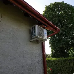 Biała jednostka zewnętrzna klimatyzatora zamontowana na ścianie domu obok brązowej rynny, widok z dołu na tle zieleni drzew i krzewów.