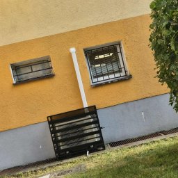 Climato Jakub Lipiński - Żółta elewacja budynku z zainstalowaną kratką wentylacyjną przy gruncie i rurą. Okna z kratami na górze. Widok z zewnątrz, zieleń trawy w tle.