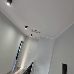 Climato Jakub Lipiński - Szary korytarz z nowoczesnym oświetleniem sufitowym i kratką wentylacyjną. Minimalistyczny design, jasne wnętrze z elementami czerni.