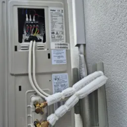 Zewnętrzna jednostka klimatyzacji Haier z otwartą skrzynką elektryczną i podłączonymi, zaizolowanymi rurami chłodniczymi, zamontowana na ścianie budynku.