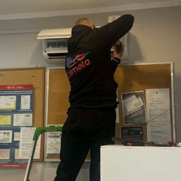 Climato Jakub Lipiński - Mężczyzna w bluzie z logo 'Climato' na drabinie, montuje klimatyzator ścienny w pomieszczeniu biurowym. Na ścianie tablice informacyjne i narzędzia.