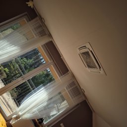 Climato Jakub Lipiński - Kasetonowa klimatyzacja sufitowa w jasnym wnętrzu, widok z dołu. Okno z firanką i grzejnik w tle. Ujęcie z perspektywy żabiej.