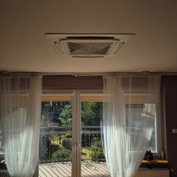 Climato Jakub Lipiński - Kasetonowa klimatyzacja sufitowa w jasnym wnętrzu z balkonem i firankami. Widok na zieleń za oknem. Promienie słoneczne podkreślają detale instalacji.