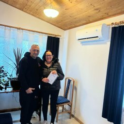 Climato Jakub Lipiński - Klimatyzator Haier zamontowany w pokoju z drewnianym sufitem. Dwie osoby stoją obok, kobieta trzyma dokumenty. Widoczne zasłony i roślina doniczkowa.