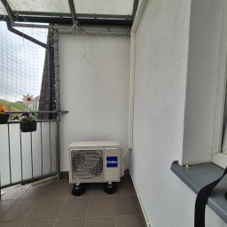 Climato Jakub Lipiński - Biała jednostka klimatyzacji Haier na balkonie z szarą podłogą i kwiatami w doniczce. Widoczne oświetlenie dekoracyjne i siatka zabezpieczająca.