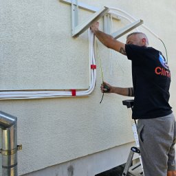 Climato Jakub Lipiński - Technik w czarnej koszulce z logo, na drabinie, precyzyjnie mierzy żółtą miarką białe rurki instalacji na elewacji budynku, pod zamontowanymi wspornikami jednostki zewnętrznej.