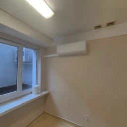 Climato Jakub Lipiński - Biała jednostka klimatyzacji ściennej w jasnym pomieszczeniu z oknem, przez które widać zewnętrzny agregat. Widoczne estetyczne rury instalacyjne i kratki wentylacyjne na suficie.