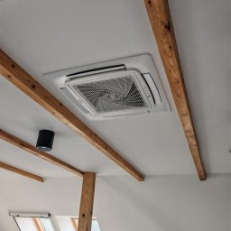 Climato Jakub Lipiński - Nowoczesny klimatyzator kasetonowy Haier w białym suficie poddasza, z drewnianymi belkami i oknami dachowymi, oświetlony naturalnym światłem.
