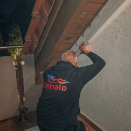Climato Jakub Lipiński - Mężczyzna w czarnej bluzie z logo Climato montuje białą osłonę instalacji na teksturowanej ścianie zewnętrznej, pod okapem dachu z dachówek, w wieczornym świetle. Widoczna metalowa balustrada.