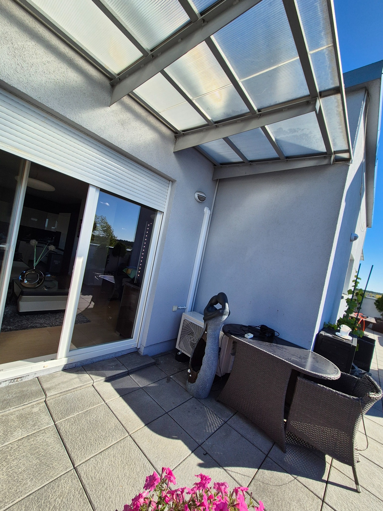 Zadaszony balkon z klimatyzatorem zewnętrznym, meblami ogrodowymi i kwiatami. Widoczne przesuwne drzwi balkonowe z roletami. Jasne płytki na podłodze i szara ściana budynku.