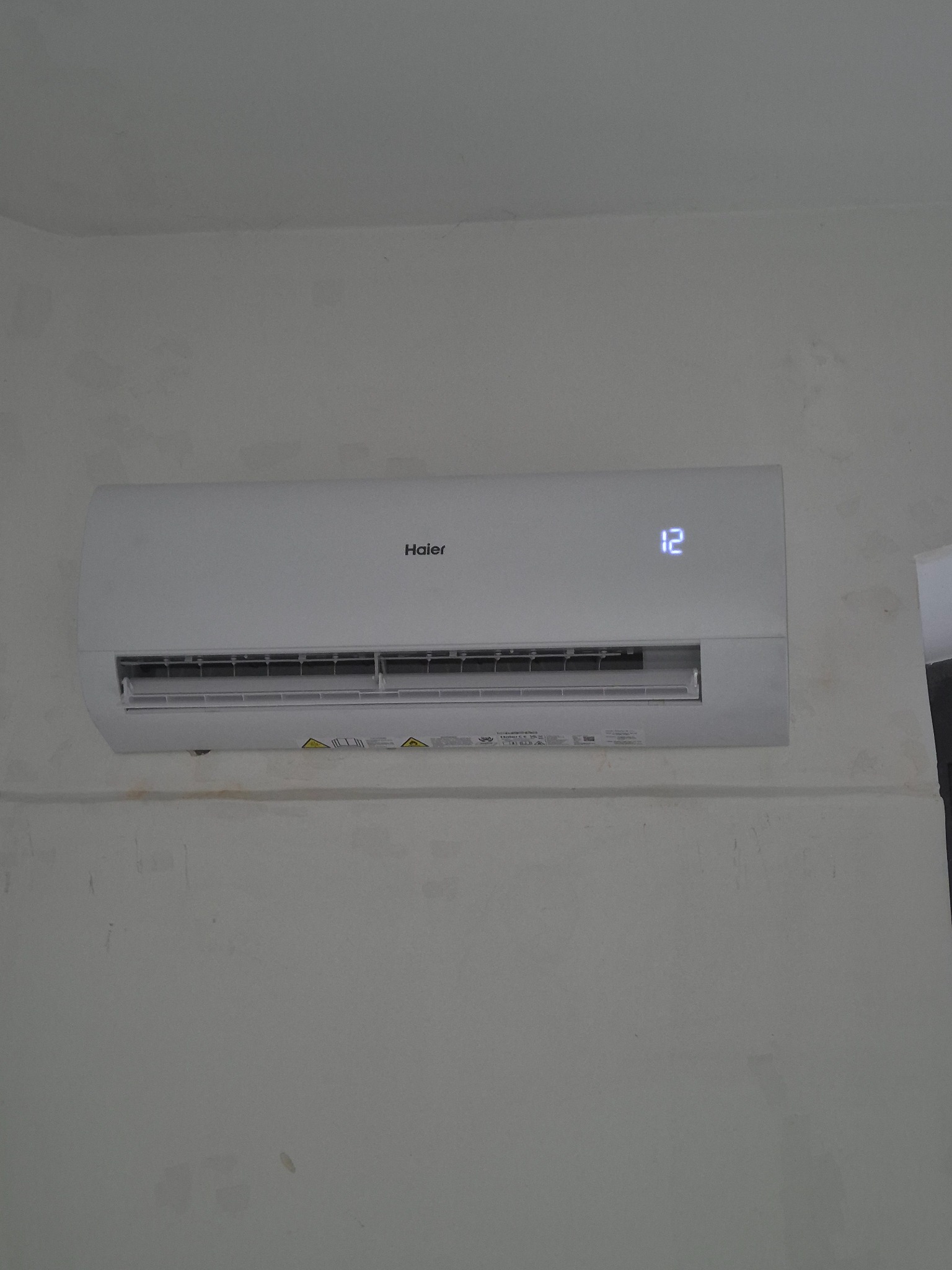 Biały klimatyzator Haier z wyświetlaczem temperatury '12' zamontowany na ścianie z widocznymi śladami po malowaniu. Widok z przodu, oświetlenie dzienne.