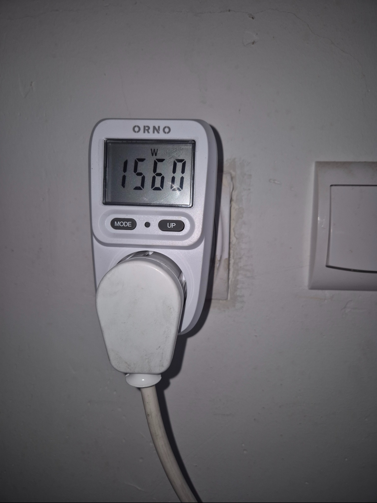 Miernik zużycia energii elektrycznej ORNO, wpięty do gniazdka, wyświetla wartość 1560 W. Obok widoczny włącznik światła na szarej ścianie.