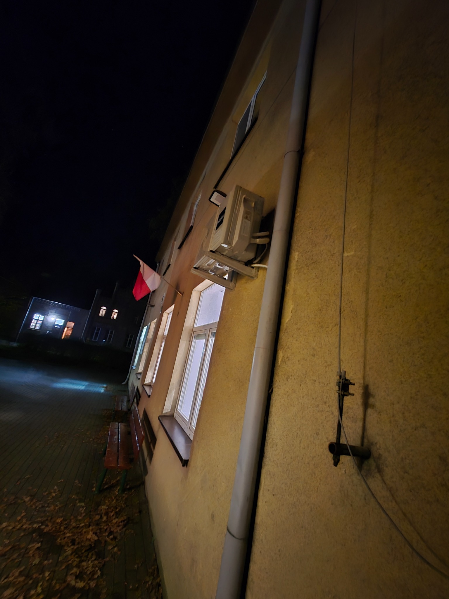 Nocny widok na budynek z klimatyzatorem na ścianie, oświetlonymi oknami i polską flagą. Ujęcie z dołu, widoczny fragment chodnika i ławki.