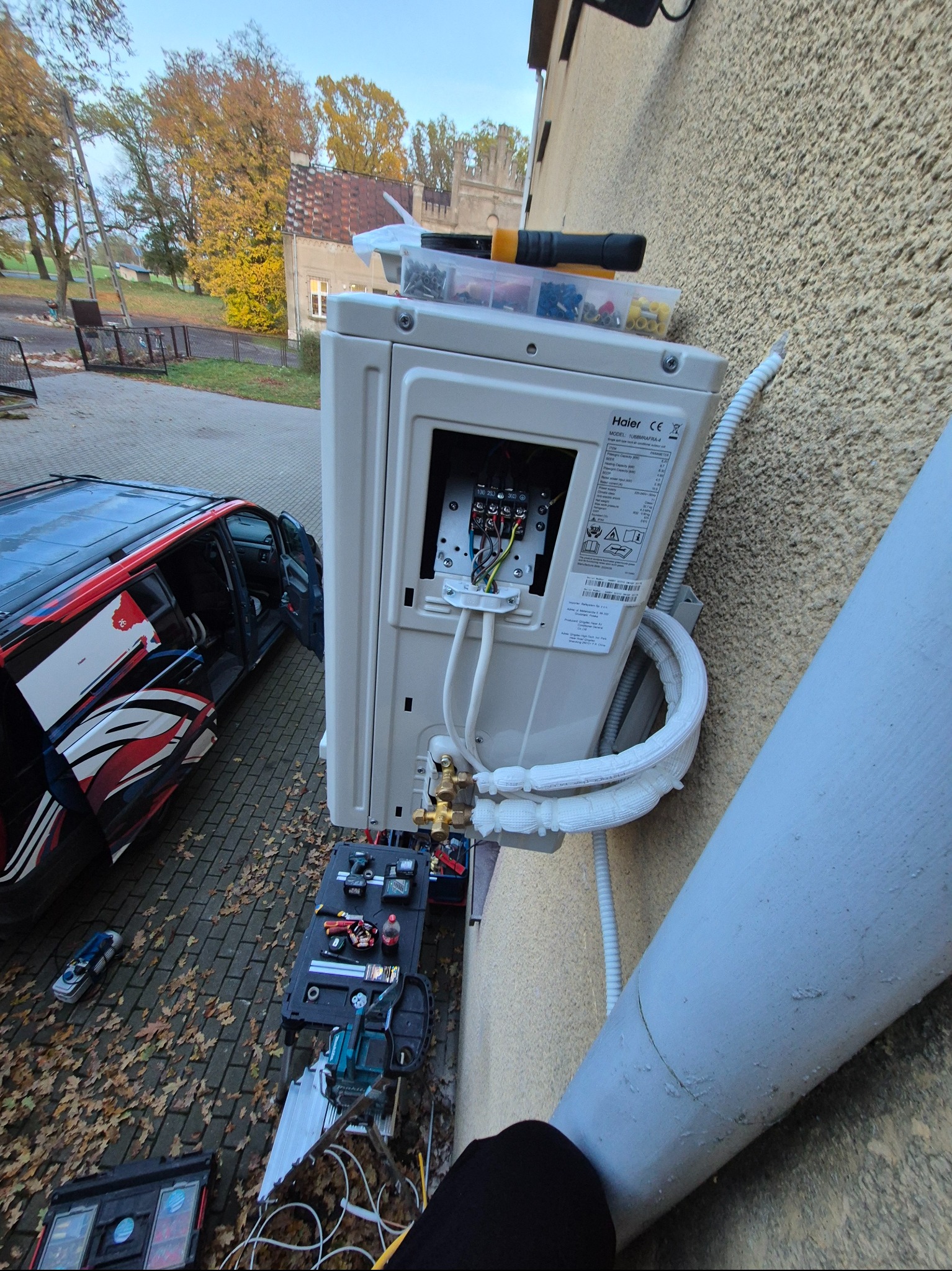Montaż klimatyzatora Haier na elewacji budynku. Widoczne narzędzia, skrzynka z elementami montażowymi i auto z otwartymi drzwiami w tle. Instalacja w trakcie podłączania.