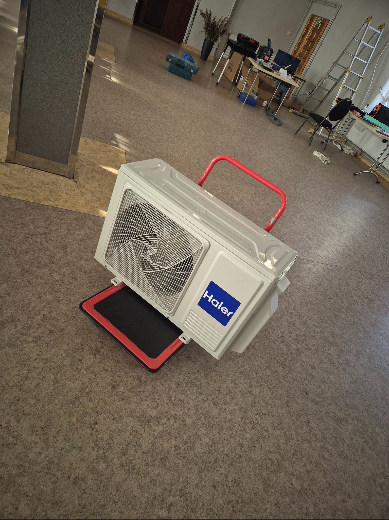 Biała jednostka zewnętrzna klimatyzacji Haier na czerwonym wózku transportowym, ustawiona ukośnie na brązowej podłodze. W tle widoczne narzędzia i drabina.