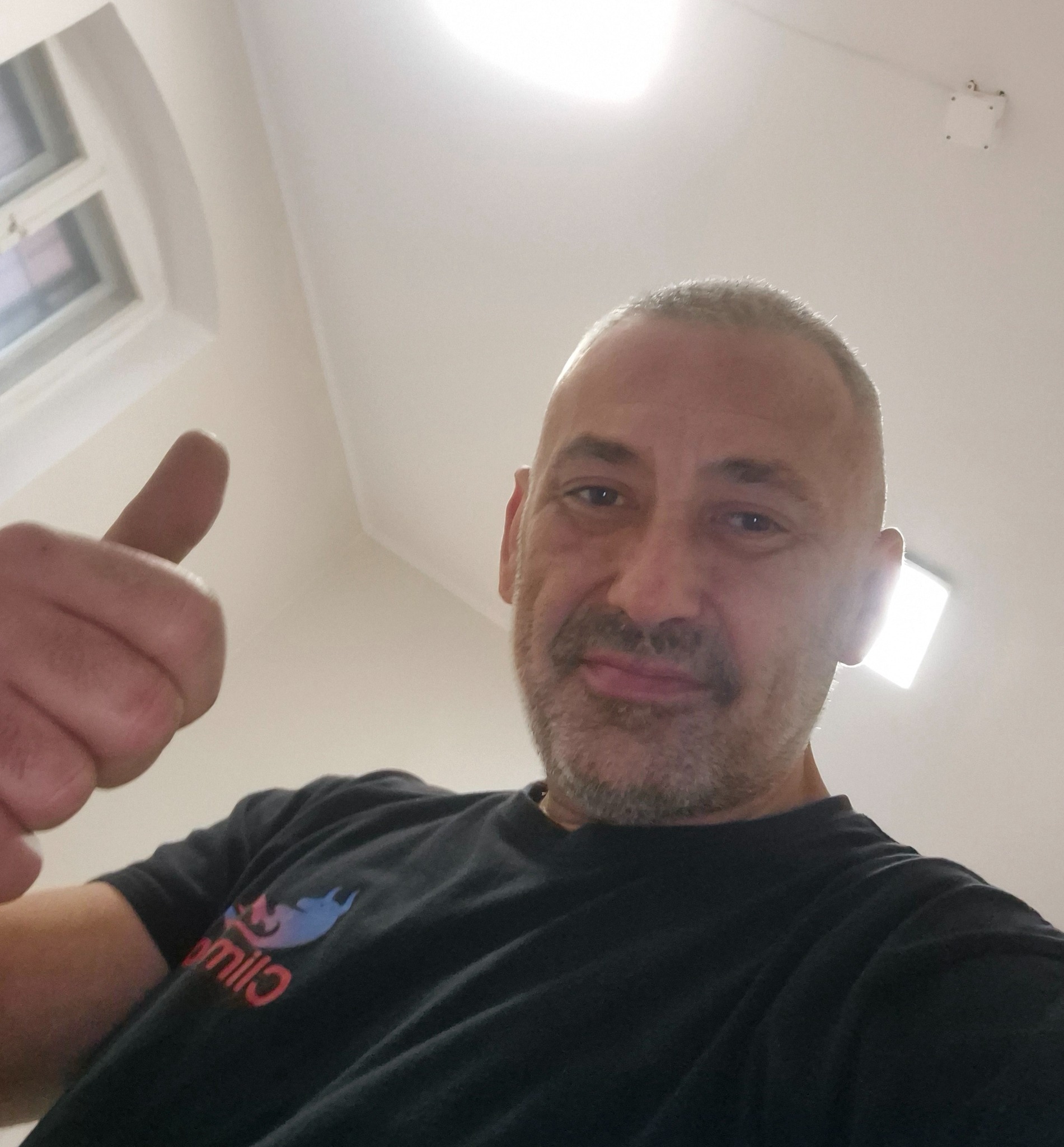 Mężczyzna z uniesionym kciukiem w górę, ubrany w czarną koszulkę z logo, na tle białego sufitu z oknem i lampą. Ujęcie z dołu.