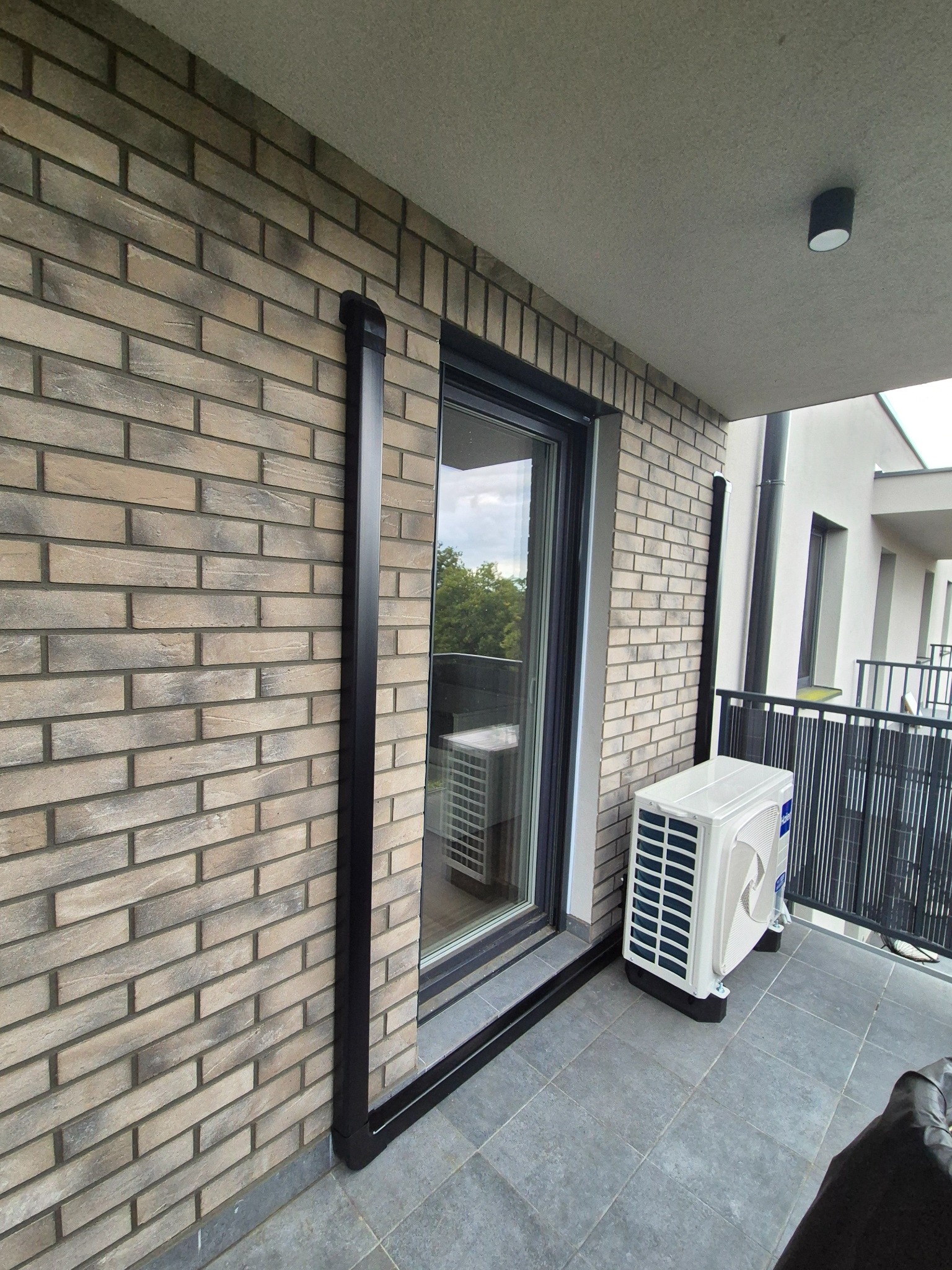 Biała jednostka zewnętrzna klimatyzacji Haier na balkonie z szarą podłogą i ceglaną ścianą. Czarna rura odprowadzająca skropliny. Widok z zewnątrz, nowoczesne budownictwo.