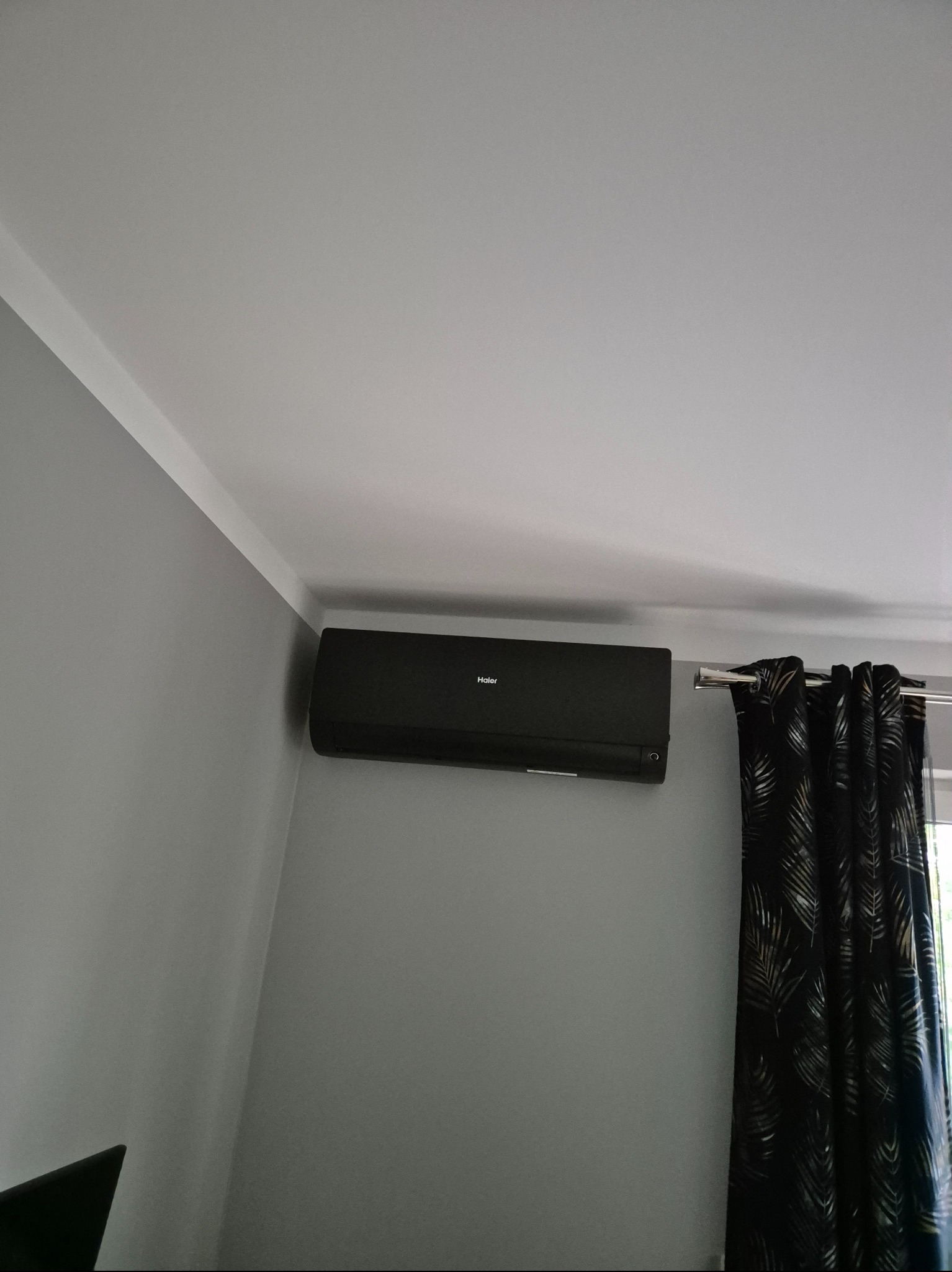 Czarny klimatyzator Haier zamontowany na szarej ścianie w pomieszczeniu z zasłonami w liście. Widok z dołu, oświetlenie naturalne.