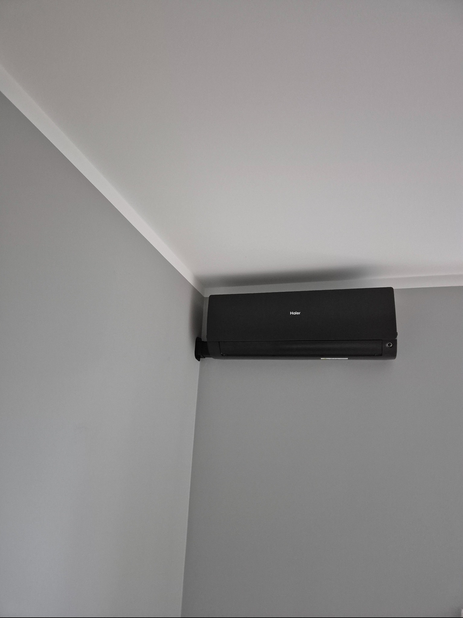 Czarny klimatyzator Haier zamontowany na szarej ścianie w rogu pomieszczenia, widoczny fragment sufitu z listwą. Minimalistyczny design.