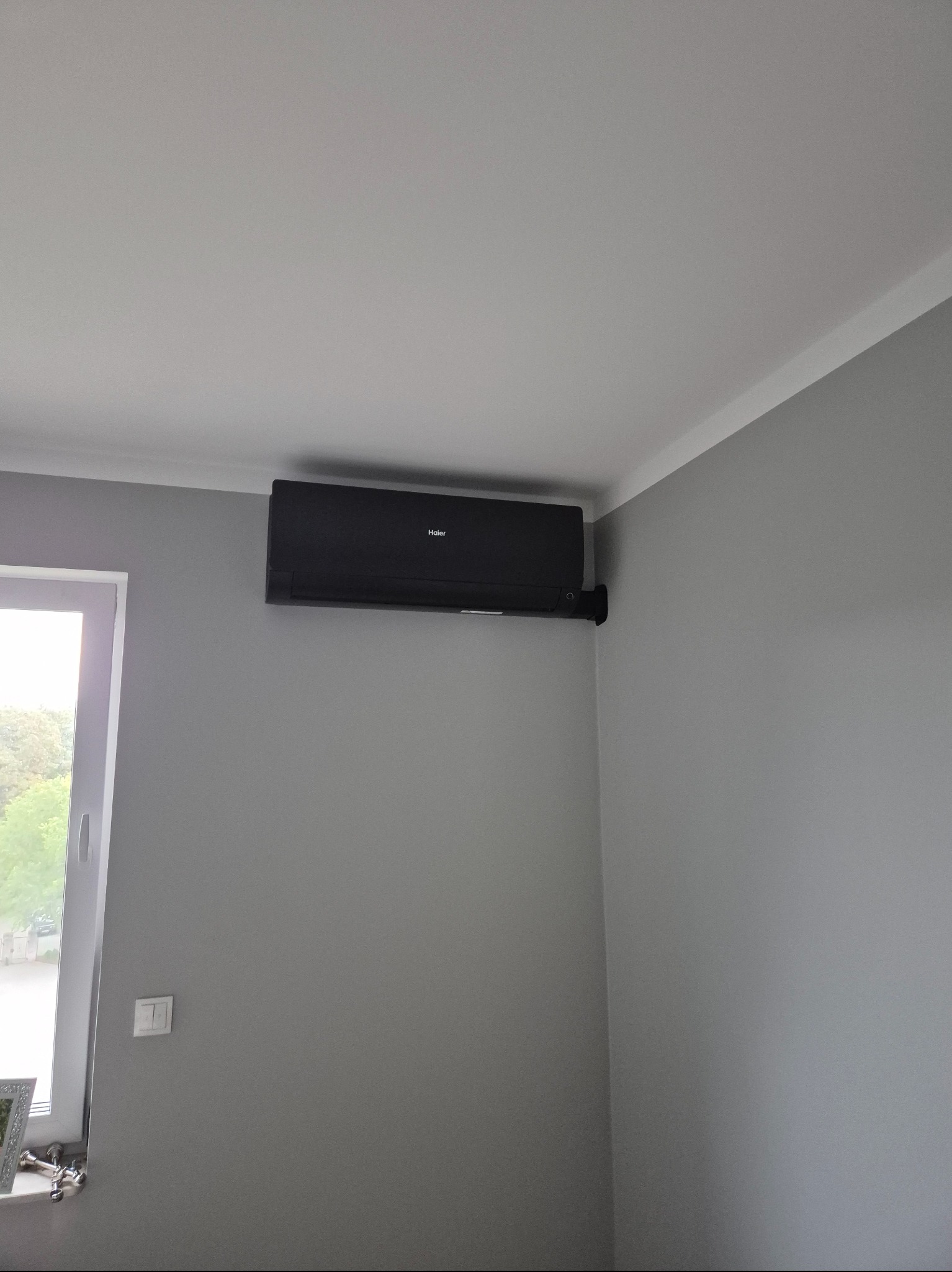 Czarny klimatyzator Haier zamontowany na szarej ścianie w rogu pokoju, widok z dołu. Obok okno z widokiem na zieleń.