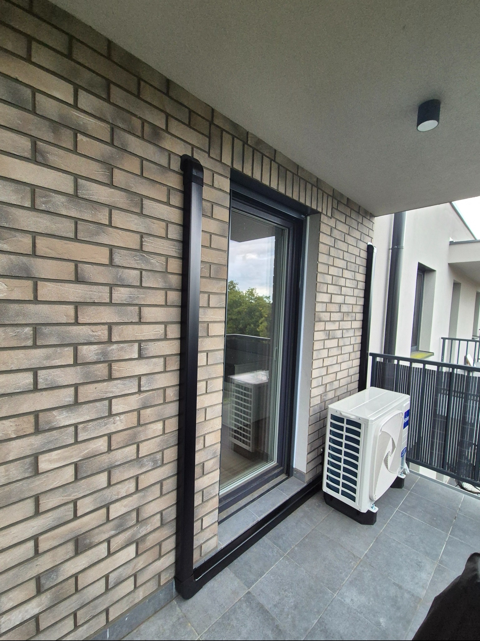 Biała jednostka zewnętrzna klimatyzacji Haier na balkonie z szarą podłogą i ceglaną ścianą. Widoczny czarny odpływ skroplin. Nowoczesne budownictwo.