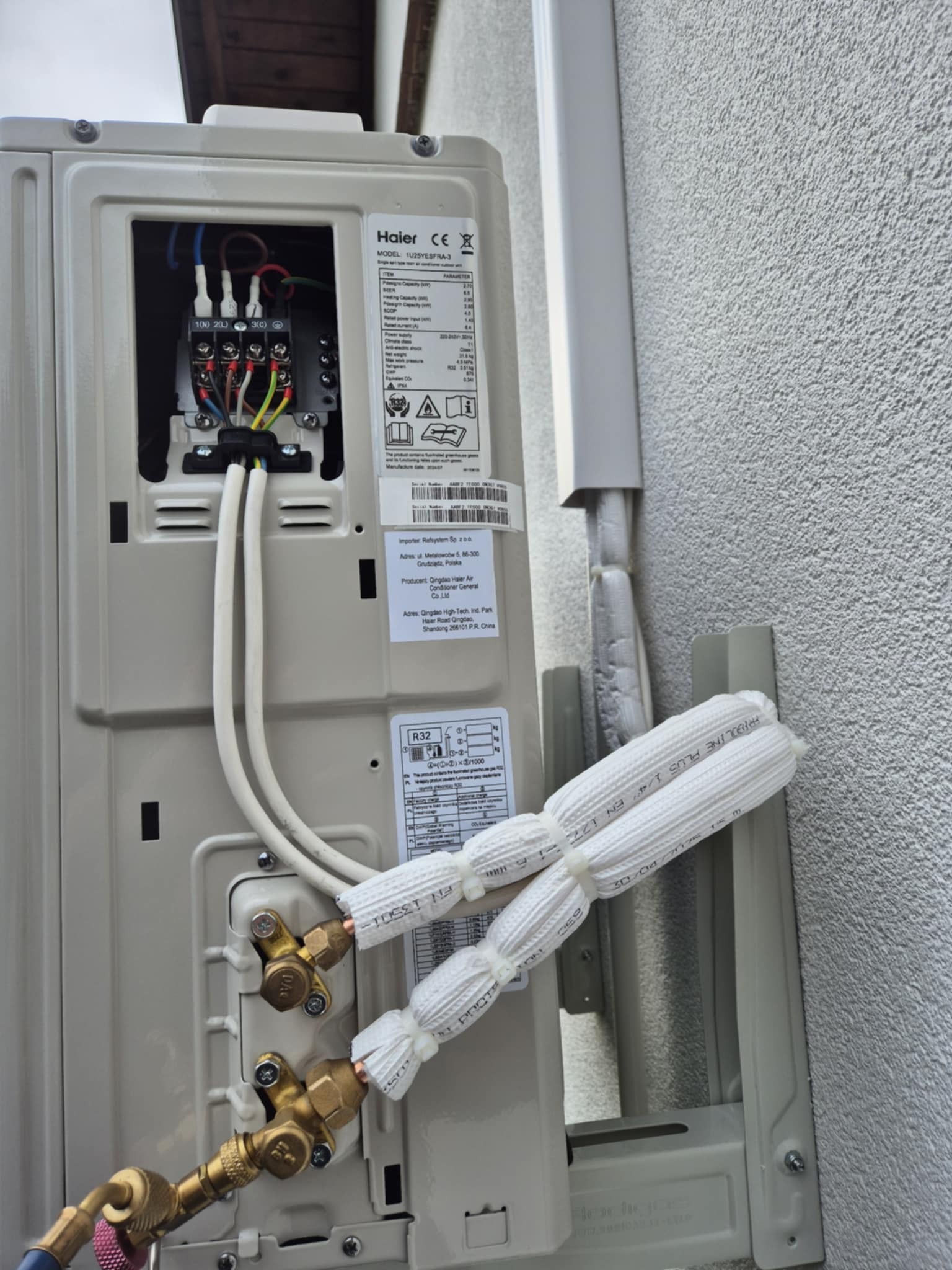 Zewnętrzna jednostka klimatyzacji Haier z otwartą skrzynką elektryczną i podłączonymi, zaizolowanymi rurami chłodniczymi, zamontowana na ścianie budynku.