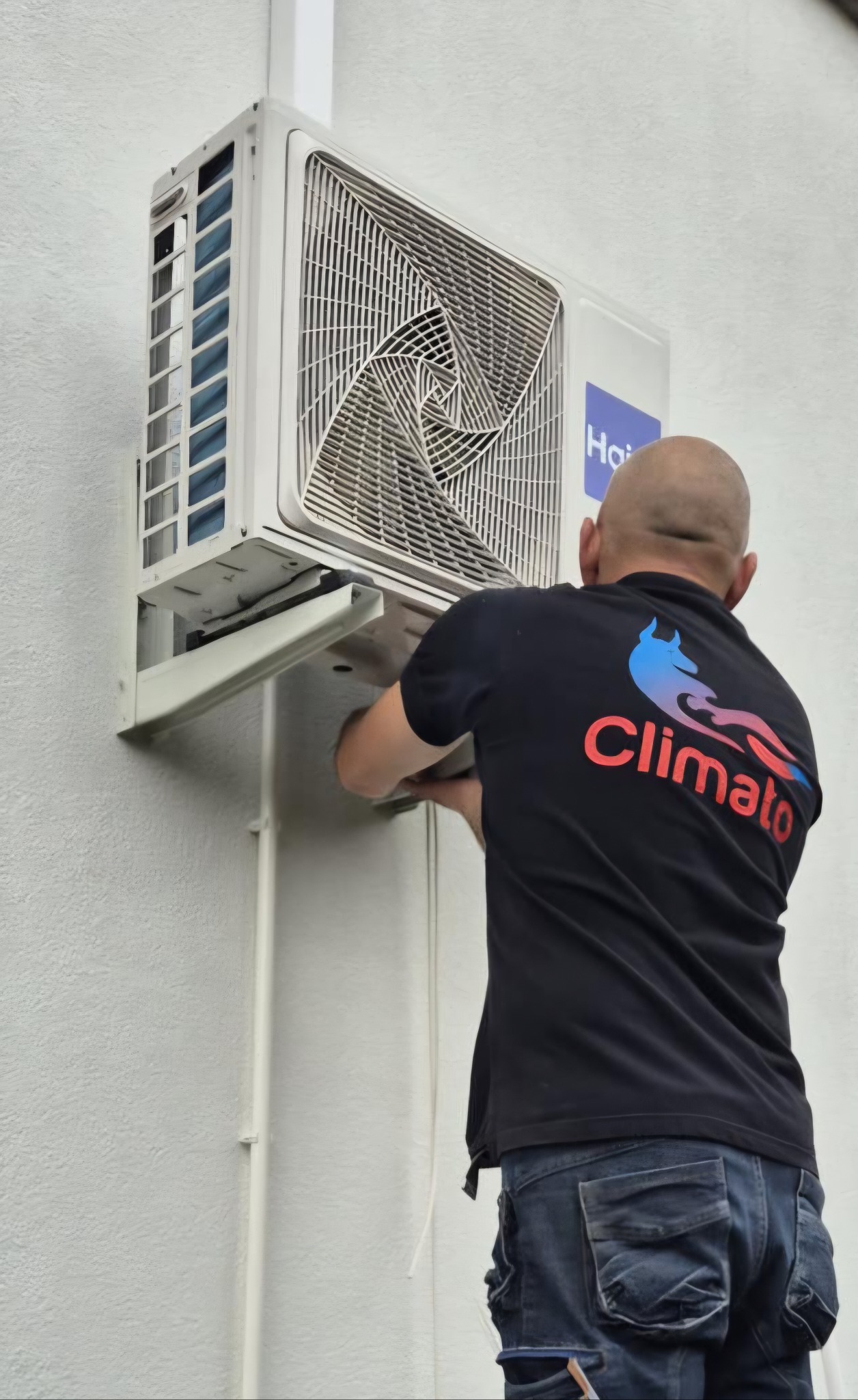 Montaż klimatyzacji Haier w Wałycz. Instalator w czarnej koszulce z logo firmy mocuje jednostkę zewnętrzną na białej ścianie budynku. Widok zza pleców.