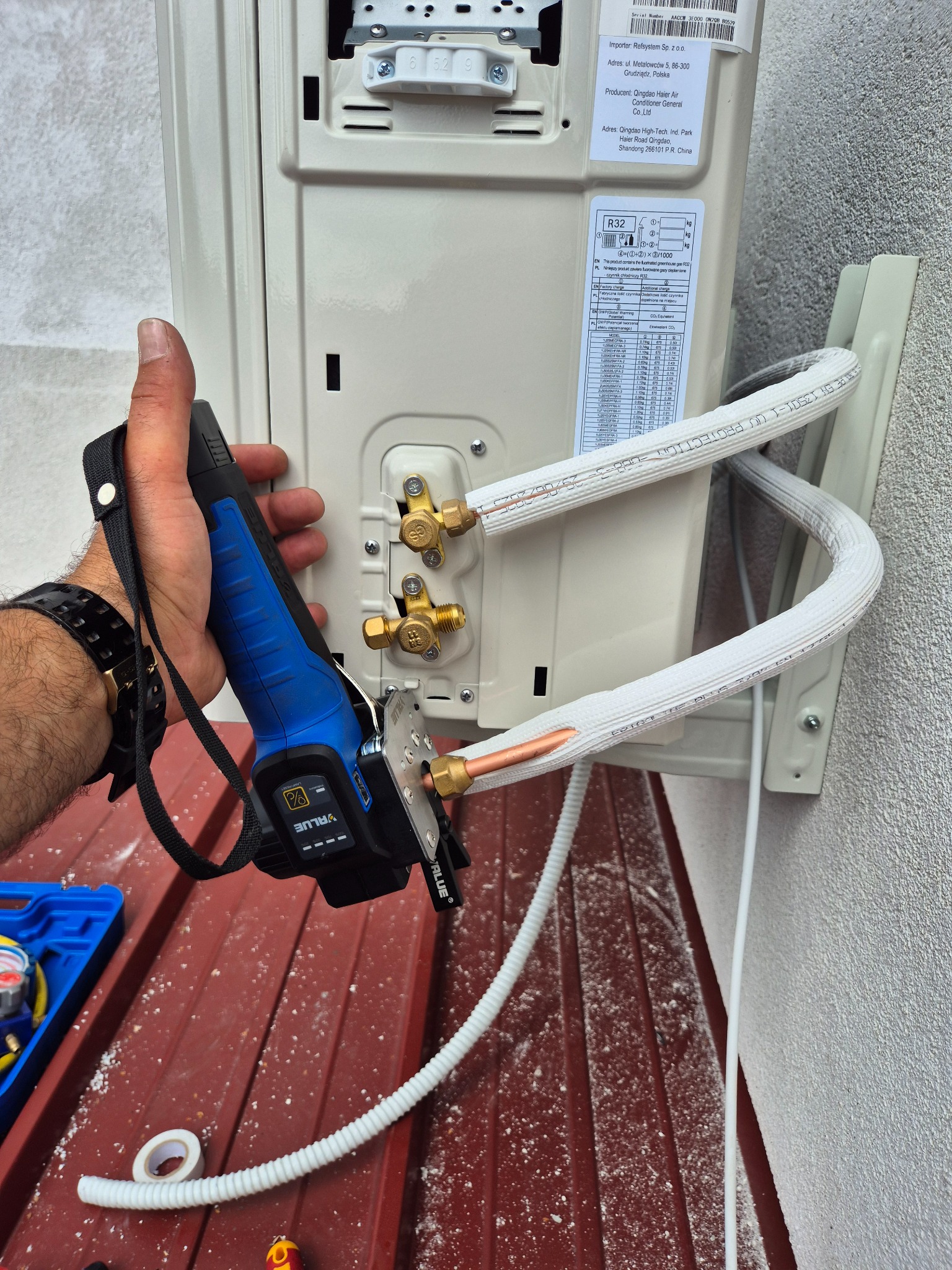 Montaż klimatyzacji: instalator używa specjalistycznego narzędzia do połączenia rur z jednostką zewnętrzną. Widoczne elementy montażowe i etykieta urządzenia.