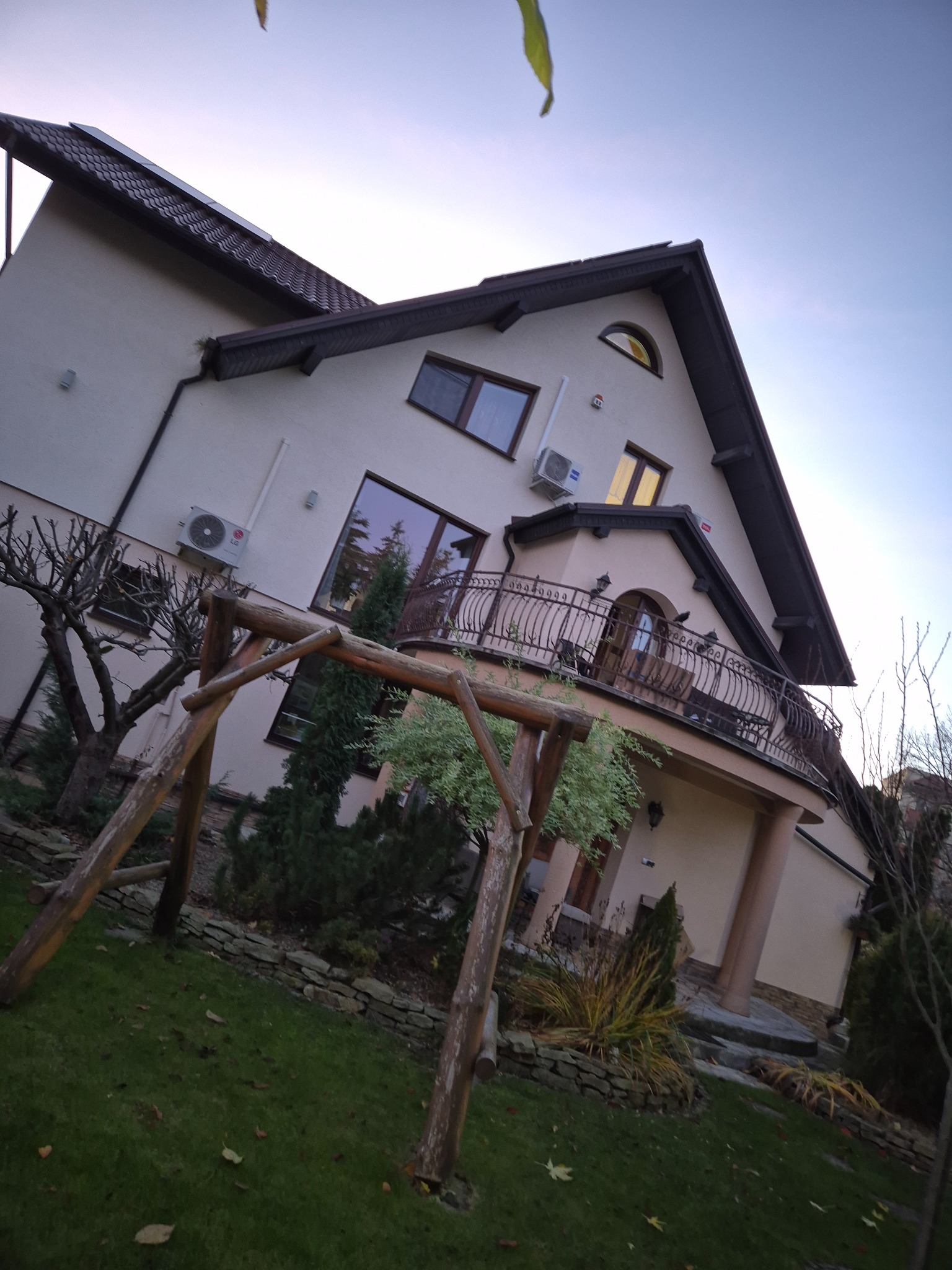 Dom jednorodzinny z balkonem i klimatyzacją na elewacji, widziany z perspektywy ogrodu z drewnianą huśtawką. Ujęcie pod kątem, wieczorne światło.