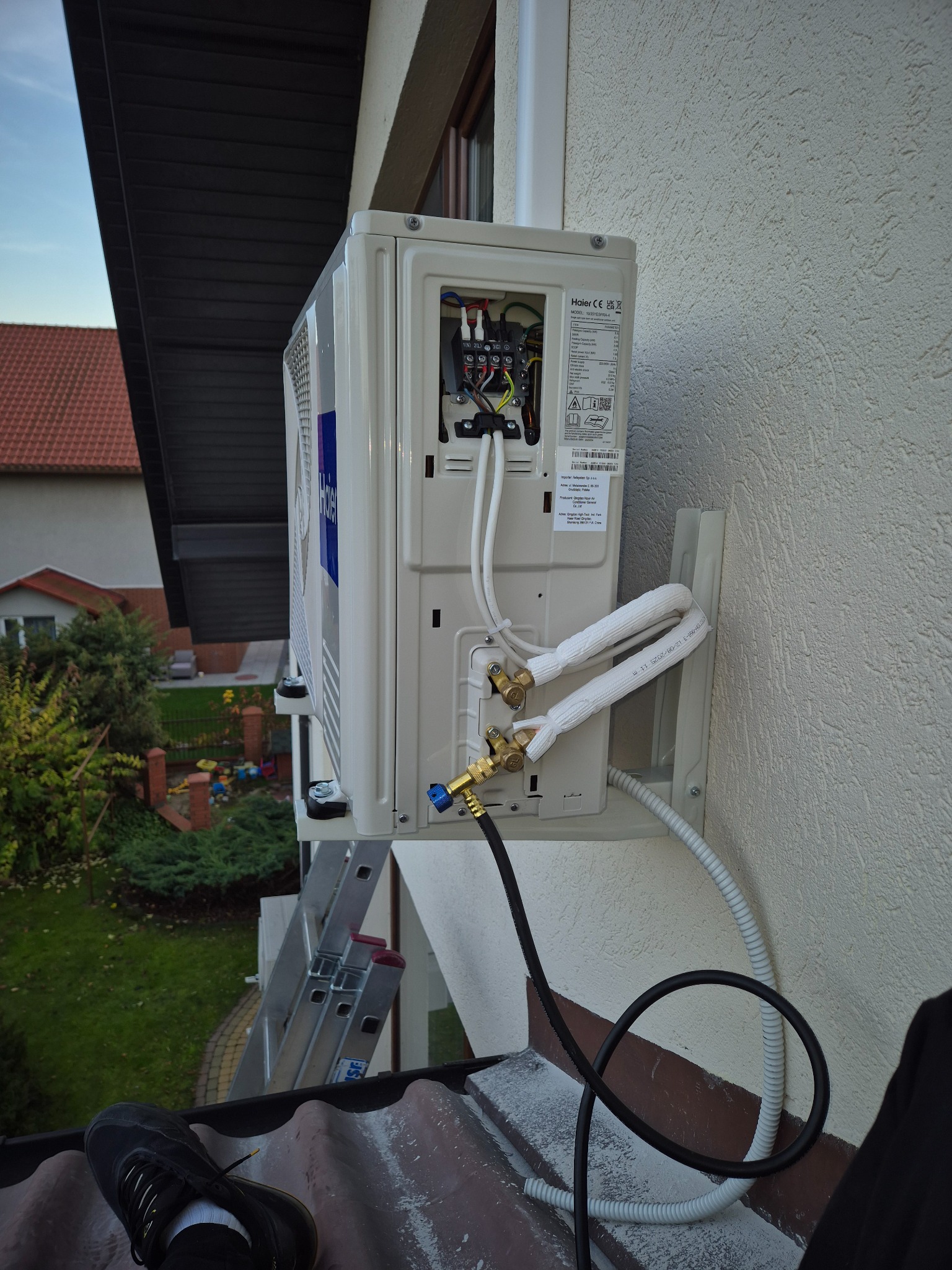Zewnętrzna jednostka klimatyzacji Haier zamontowana na ścianie budynku, widoczne podłączenia przewodów i instalacja elektryczna. Częściowo widoczna drabina i otoczenie.