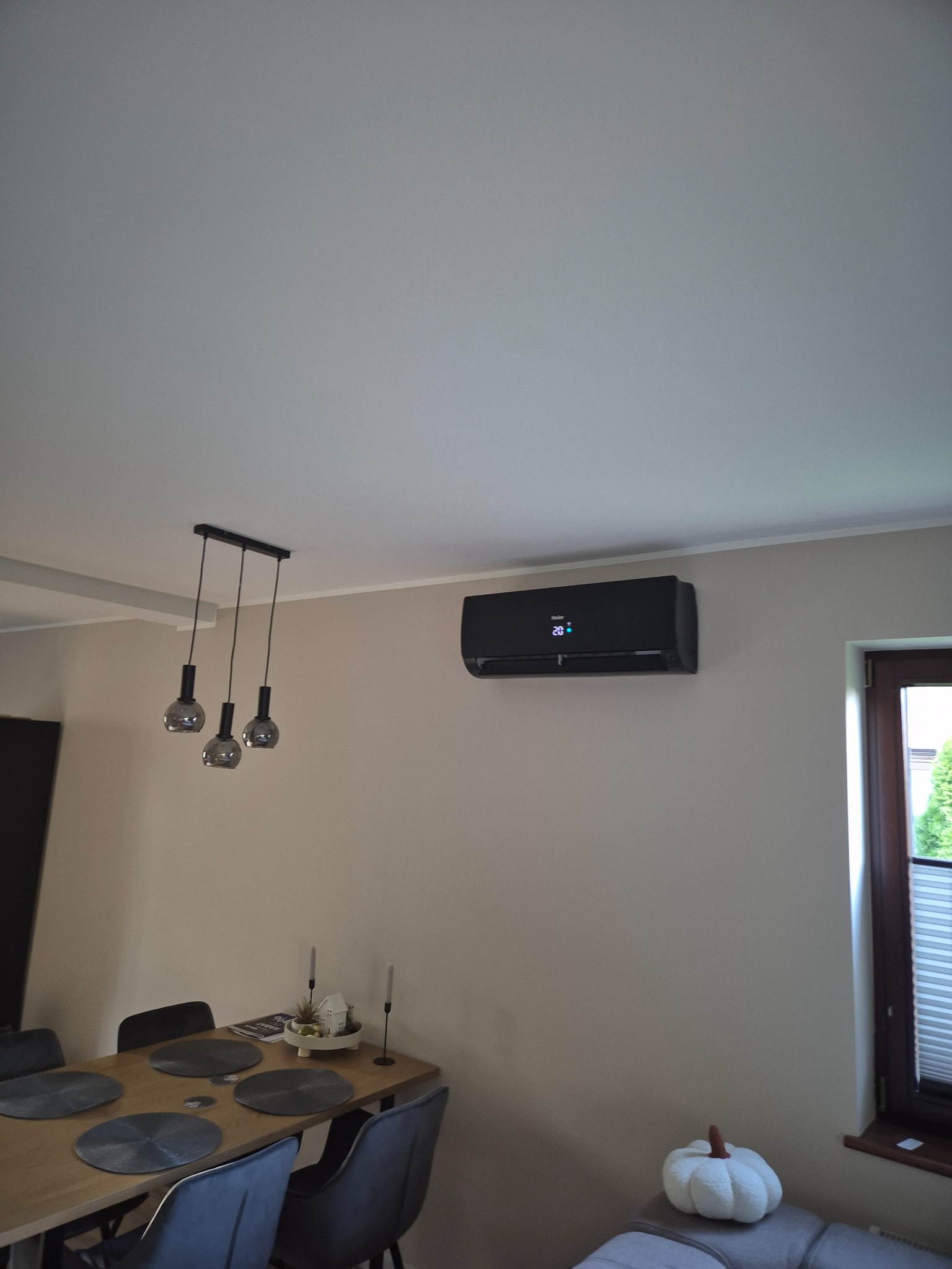 Czarny klimatyzator Haier zamontowany na ścianie w jasnym wnętrzu, widoczny stół z krzesłami i lampa wisząca. Nowoczesny design, estetyczny montaż.